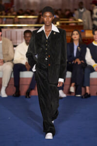 Model walking a fashion runway in a black pinstripe suit with a white shirt collar; audience seated in the background.