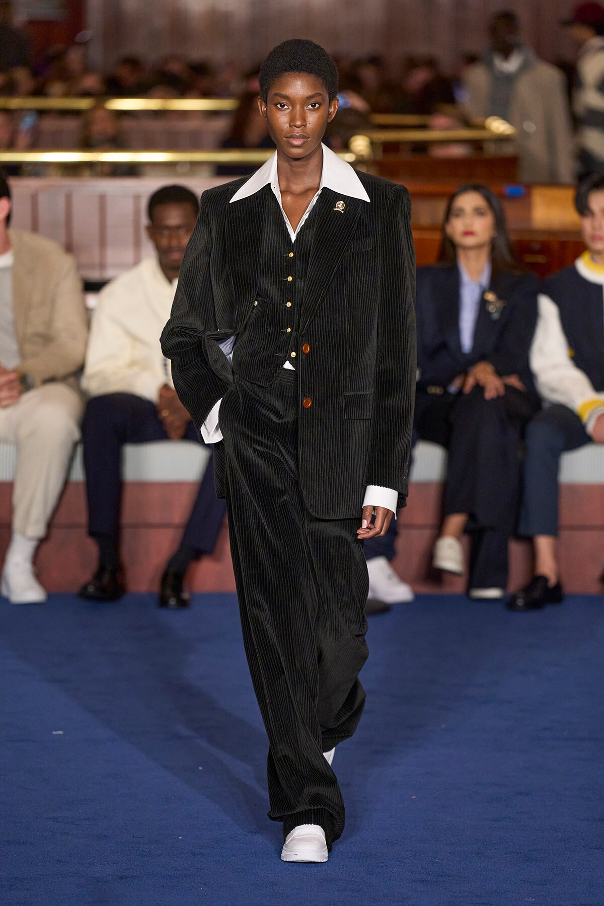 Model walking a fashion runway in a black pinstripe suit with a white shirt collar; audience seated in the background.