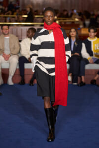 Model walking runway in black-and-white striped shirt, black shorts, knee-high boots, and a bright red scarf; blurred audience in background.