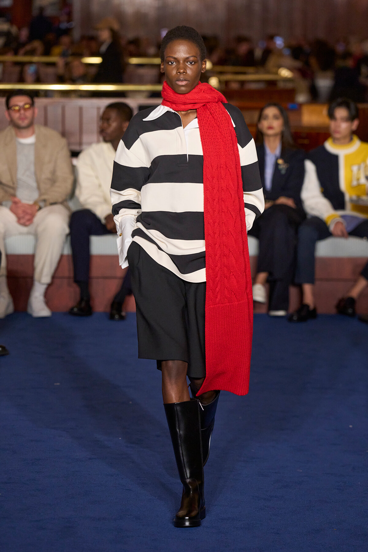 Model walking runway in black-and-white striped shirt, black shorts, knee-high boots, and a bright red scarf; blurred audience in background.