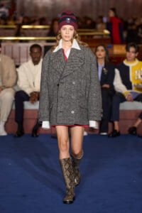 Model walks a runway in a oversized grey herringbone double-breasted coat, red trim, white shirt cuffs, burgundy beanie, and snakeskin knee-high boots.
