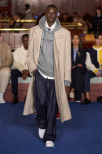 Male model walks the runway in a beige overcoat layered over a gray hoodie and navy pinstripe pants with white sneakers.