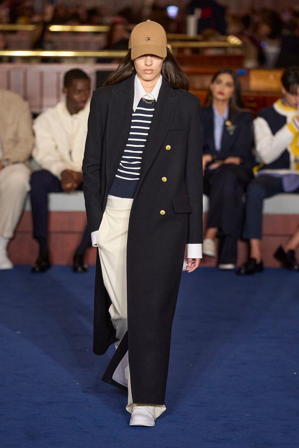 Model walks a fashion runway in a long black double-breasted coat with gold buttons, striped navy sweater, cream wide-leg pants, white sneakers, and a tan cap.