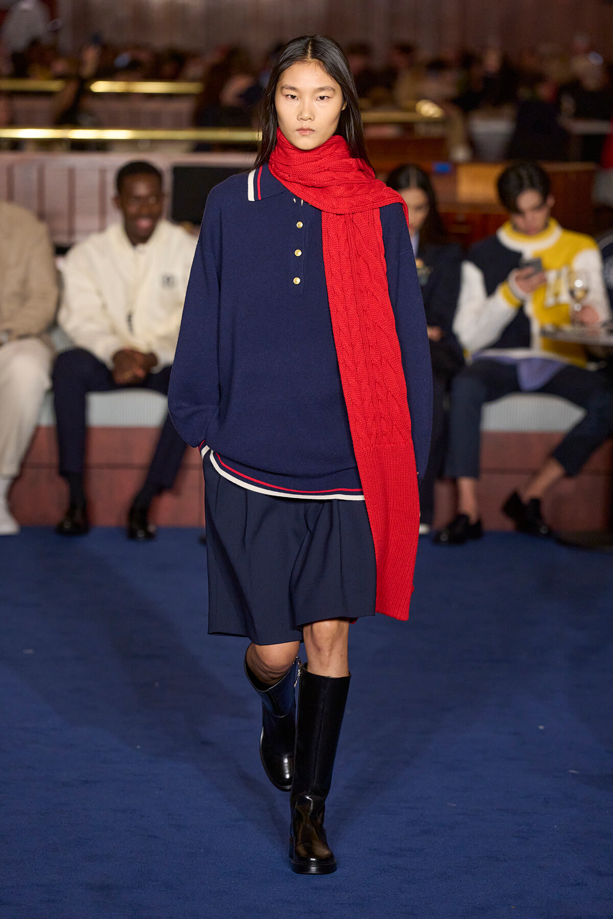 Model walks a runway in a navy outfit with a bright red scarf, knee-high black boots, and a layered navy skirt; audience sits along the sides in a fashion show setting.