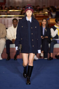 Model walking a fashion show in a navy double-breasted coat with gold buttons, burgundy knit hat, and knee-high boots, carrying a small black bag
