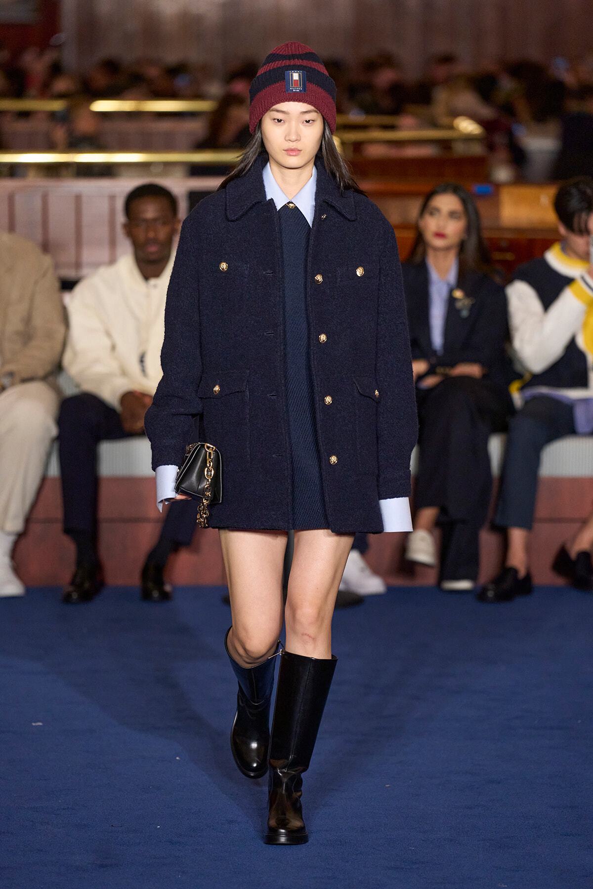 Model walking a fashion show in a navy double-breasted coat with gold buttons, burgundy knit hat, and knee-high boots, carrying a small black bag