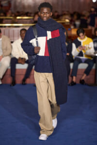 Male model walks the runway in an oversized navy color-block knit sweater, beige trousers, white sneakers, and a navy scarf with a shoulder bag.