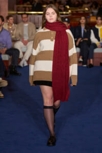 Models a red scarf and oversized striped sweater as she walks a fashion runway with audience in the background.