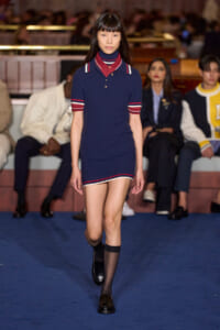 Runway model walks in a navy knit polo dress with red and white trim, black shoes, on a blue catwalk with seated audience in the background.