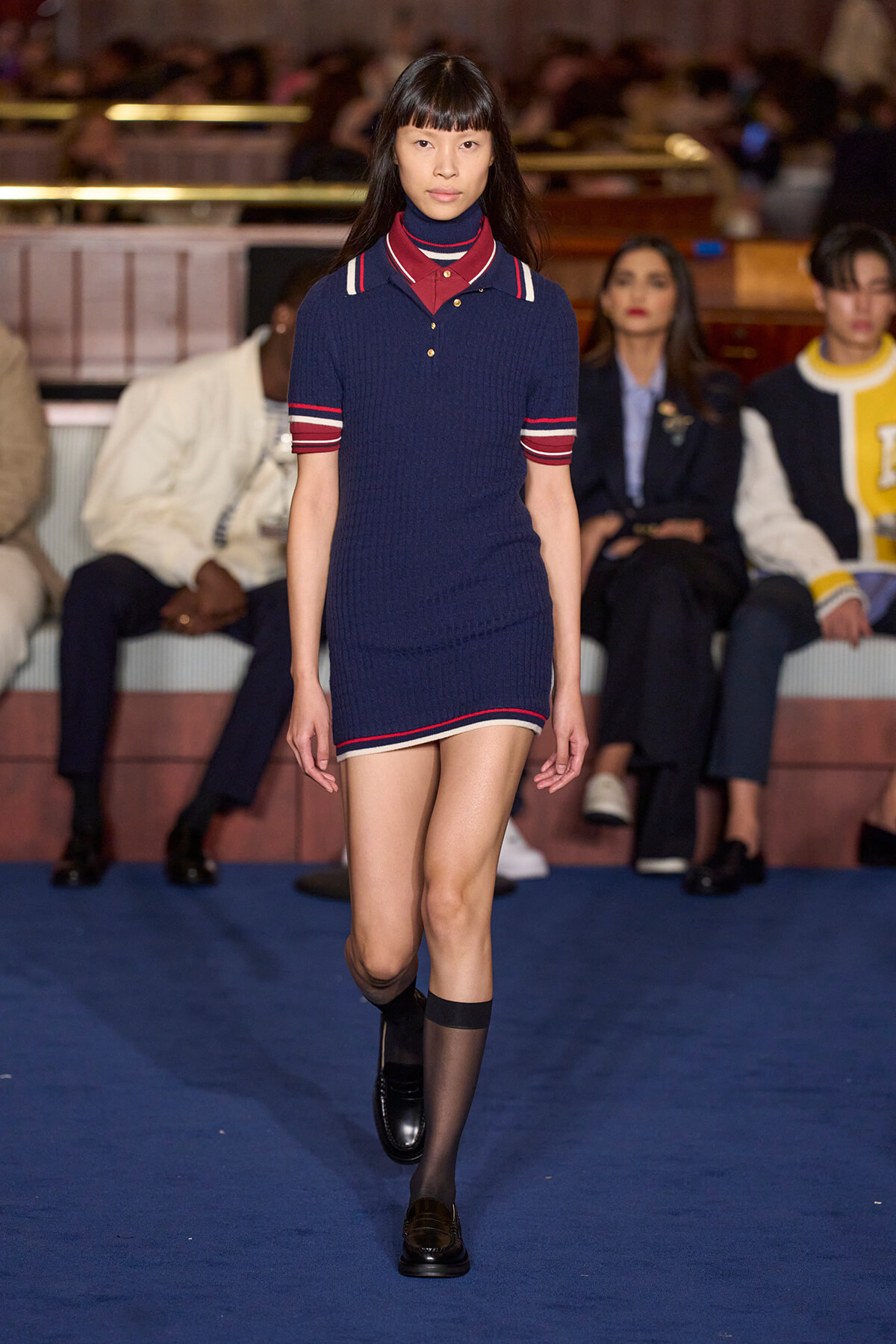 Runway model walks in a navy knit polo dress with red and white trim, black shoes, on a blue catwalk with seated audience in the background.