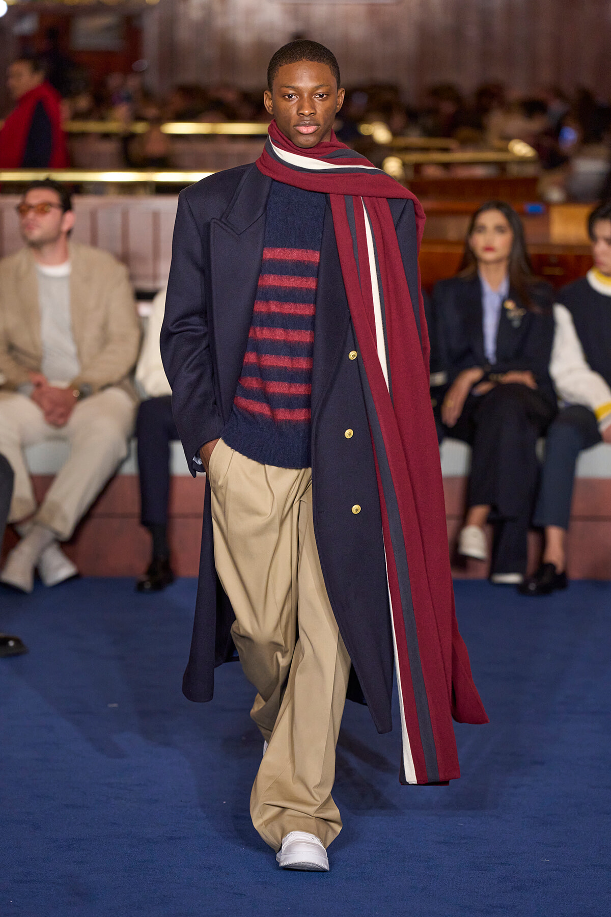 Male model walking a fashion show runway in a navy coat, maroon scarf, striped navy sweater, and tan trousers; audience blurred in background.