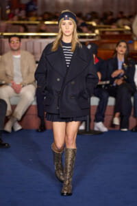 Model walks the runway in a navy oversized coat, striped top, beanie, and snakeskin boots on a blue catwalk.