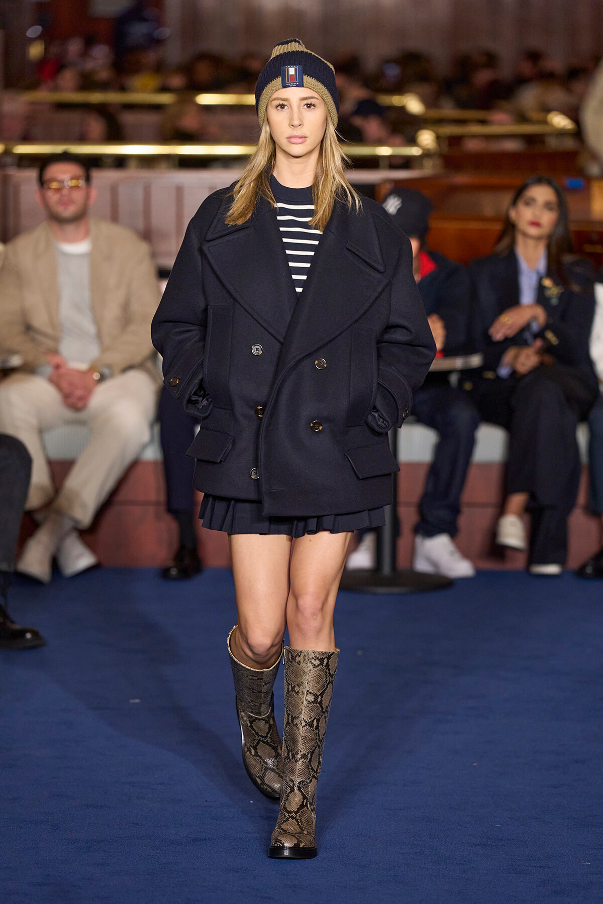 Model walks the runway in a navy oversized coat, striped top, beanie, and snakeskin boots on a blue catwalk.