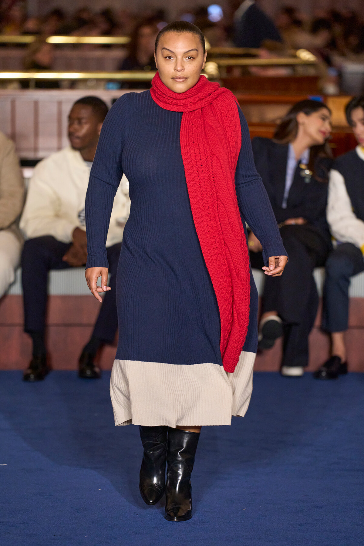 Model walking a runway in a navy knit dress with a red scarf and beige pleated hem, black knee-high boots, audience in background