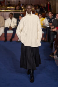 Model walks the runway in an oversized cream cable-knit turtleneck sweater and a long black skirt on a blue catwalk with spectators in the background.