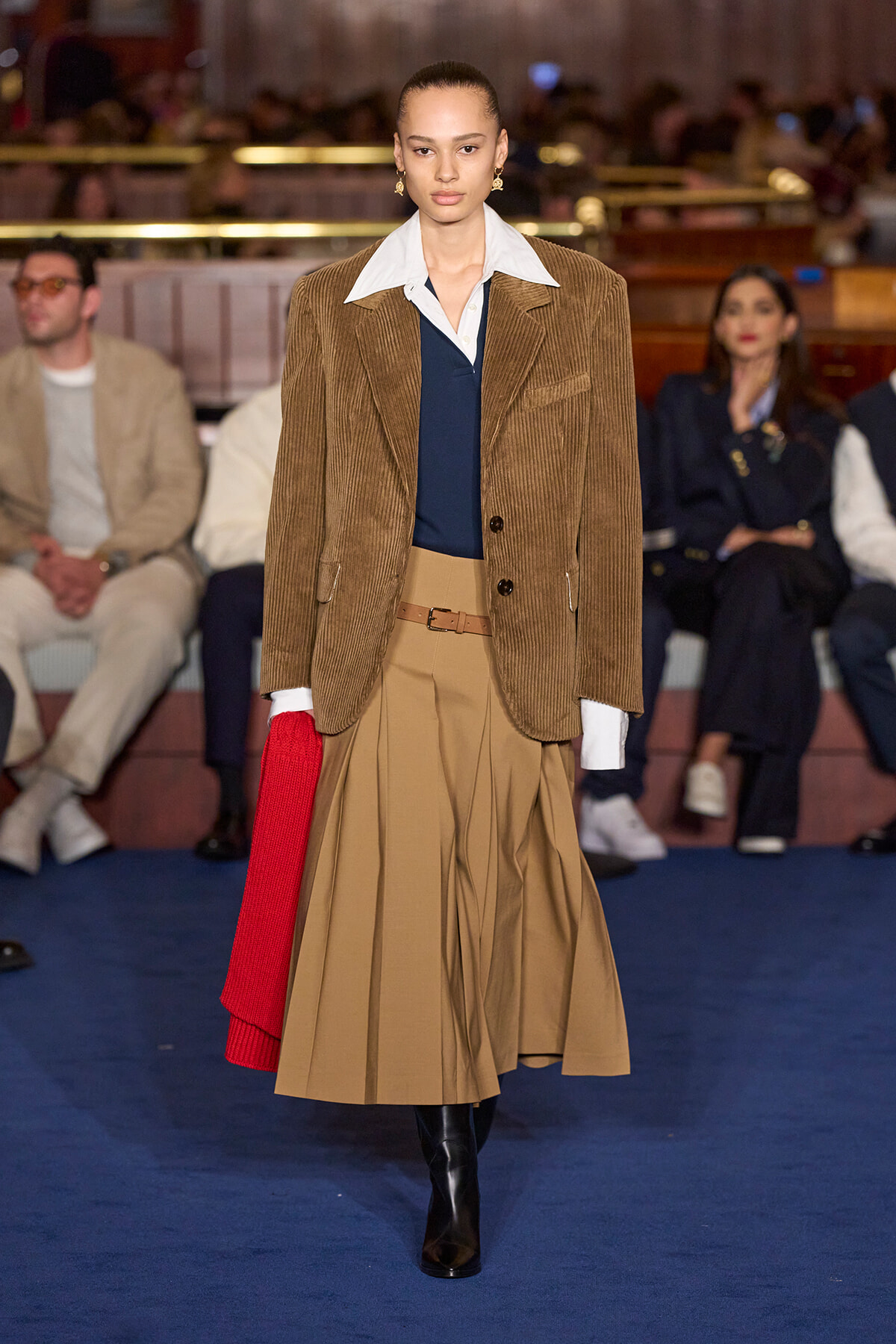 Model on a runway wearing a brown corduroy blazer over a white-collar shirt, navy top, and a tan pleated midi skirt with black boots and a red scarf draped at the side.