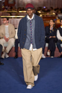 Male model walking the runway in a navy knit cardigan over a checkered shirt and striped tie, beige trousers, white sneakers, and a red cap.