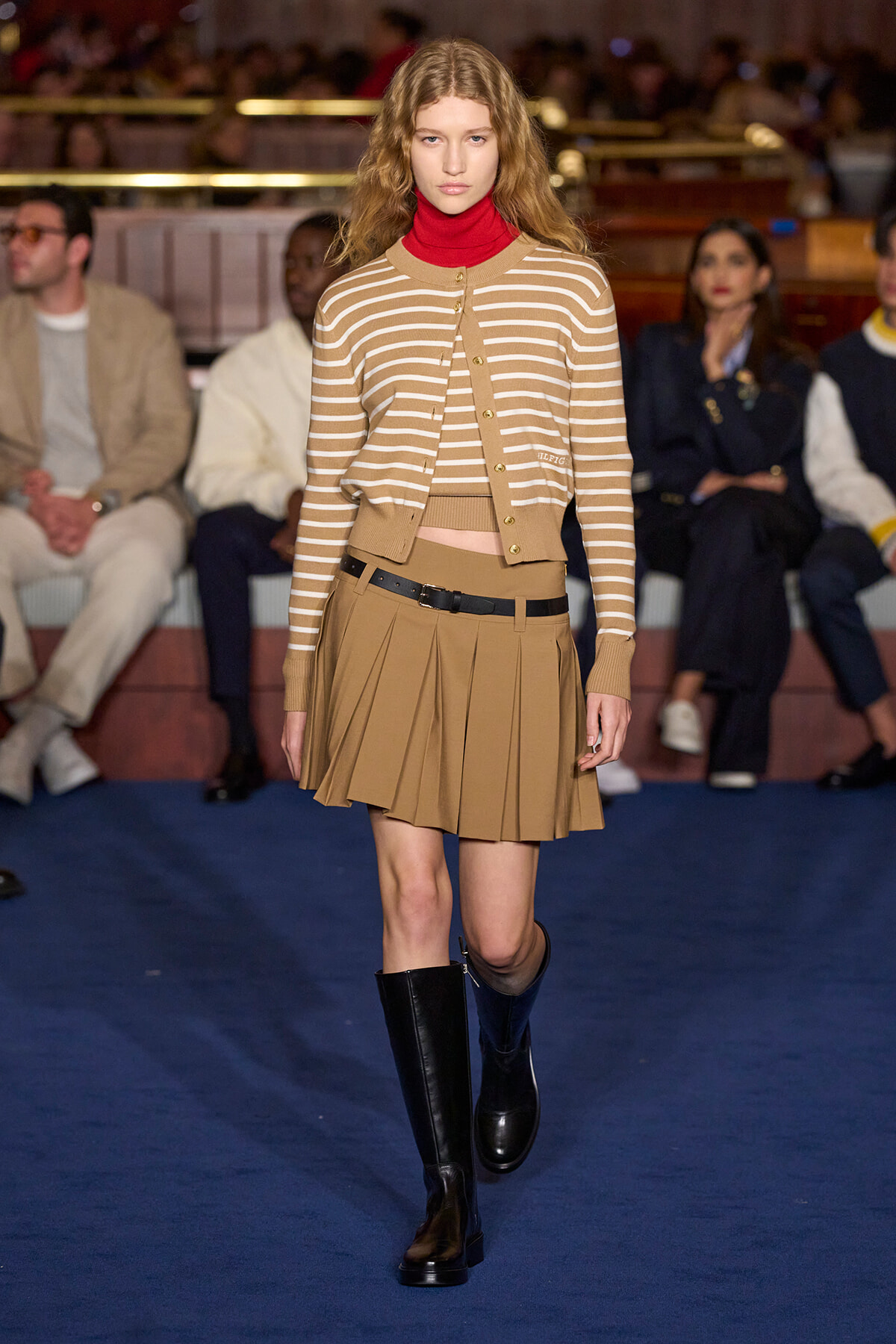 Runway model walking the catwalk in a camel-striped cardigan over a red turtleneck and pleated tan skirt with a black belt and knee-high boots.