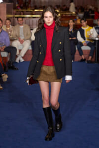 Model walks the runway in a red turtleneck under a black double-breasted blazer with gold buttons, a brown mini skirt, and knee-high black boots, audience in background.