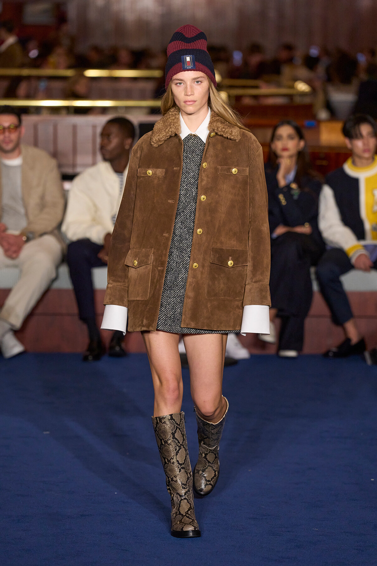 Model walking a runway in a brown corduroy jacket with shearling collar, gray dress, and snake-print boots; audience blurred in background.
