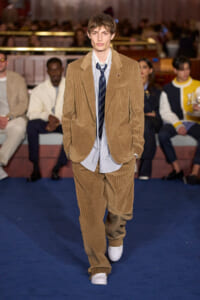 Male model walking a fashion show runway in a tan corduroy suit with a striped shirt and navy tie, white sneakers, audience in the background.