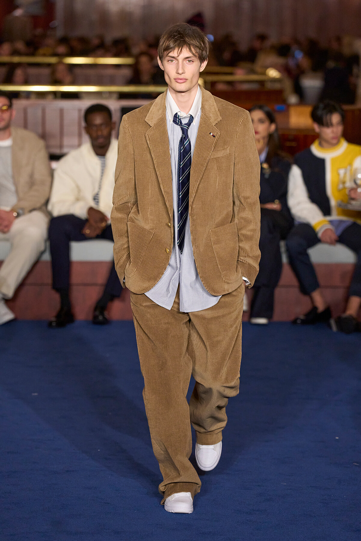 Male model walking a fashion show runway in a tan corduroy suit with a striped shirt and navy tie, white sneakers, audience in the background.