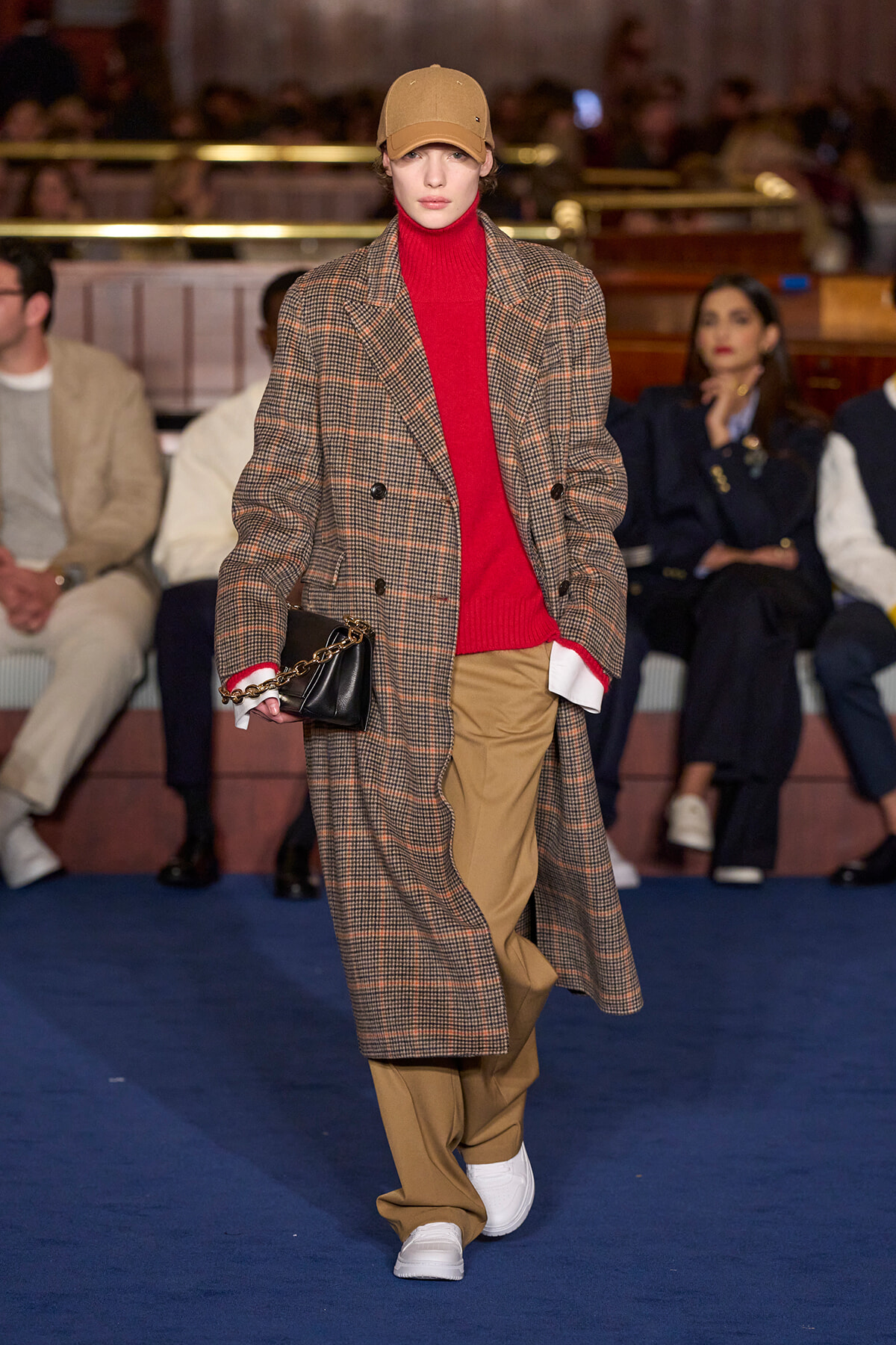 Model walking a runway in a brown plaid double-breasted coat over a red turtleneck, tan trousers, beige cap, white sneakers, and a black chain-strap bag.