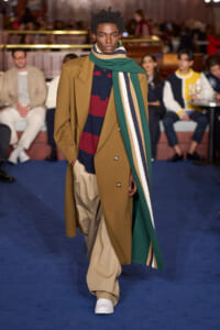 Male model walks the runway in a camel coat, navy-red striped sweater, tan trousers, and a long green striped scarf.