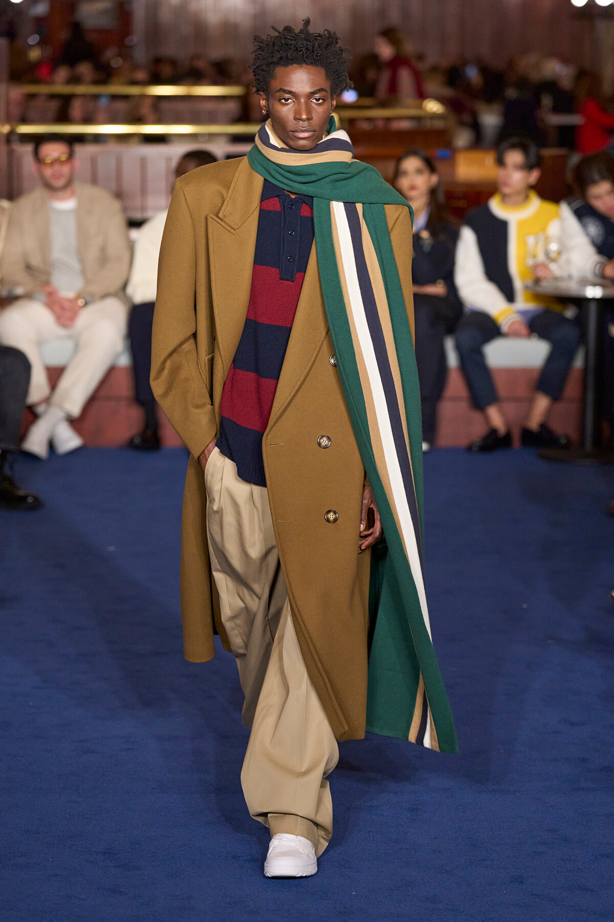 Male model walks the runway in a camel coat, navy-red striped sweater, tan trousers, and a long green striped scarf.