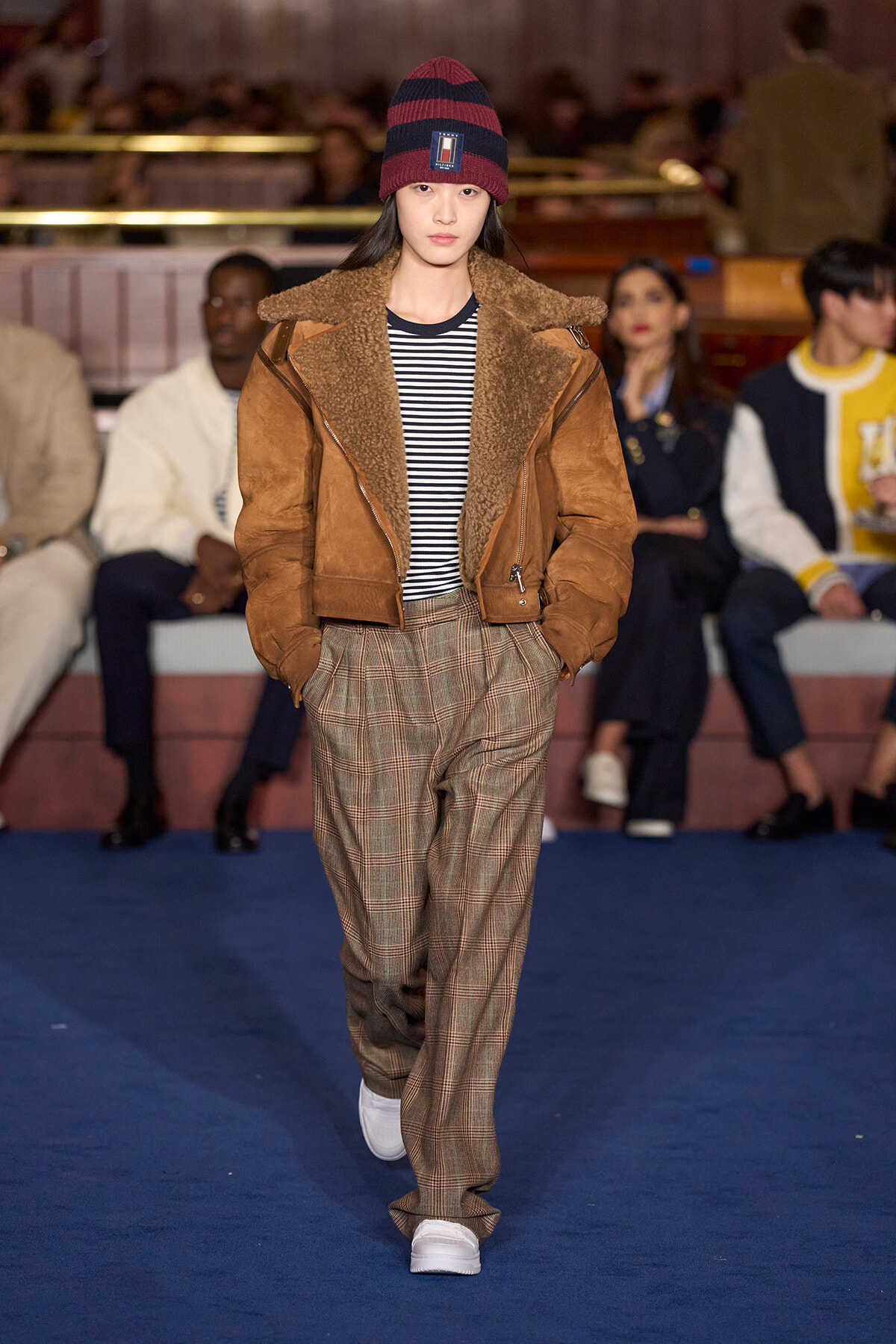 Model walking a fashion runway in a brown shearling jacket, striped shirt, plaid trousers, white sneakers, and a red-and-navy beanie.