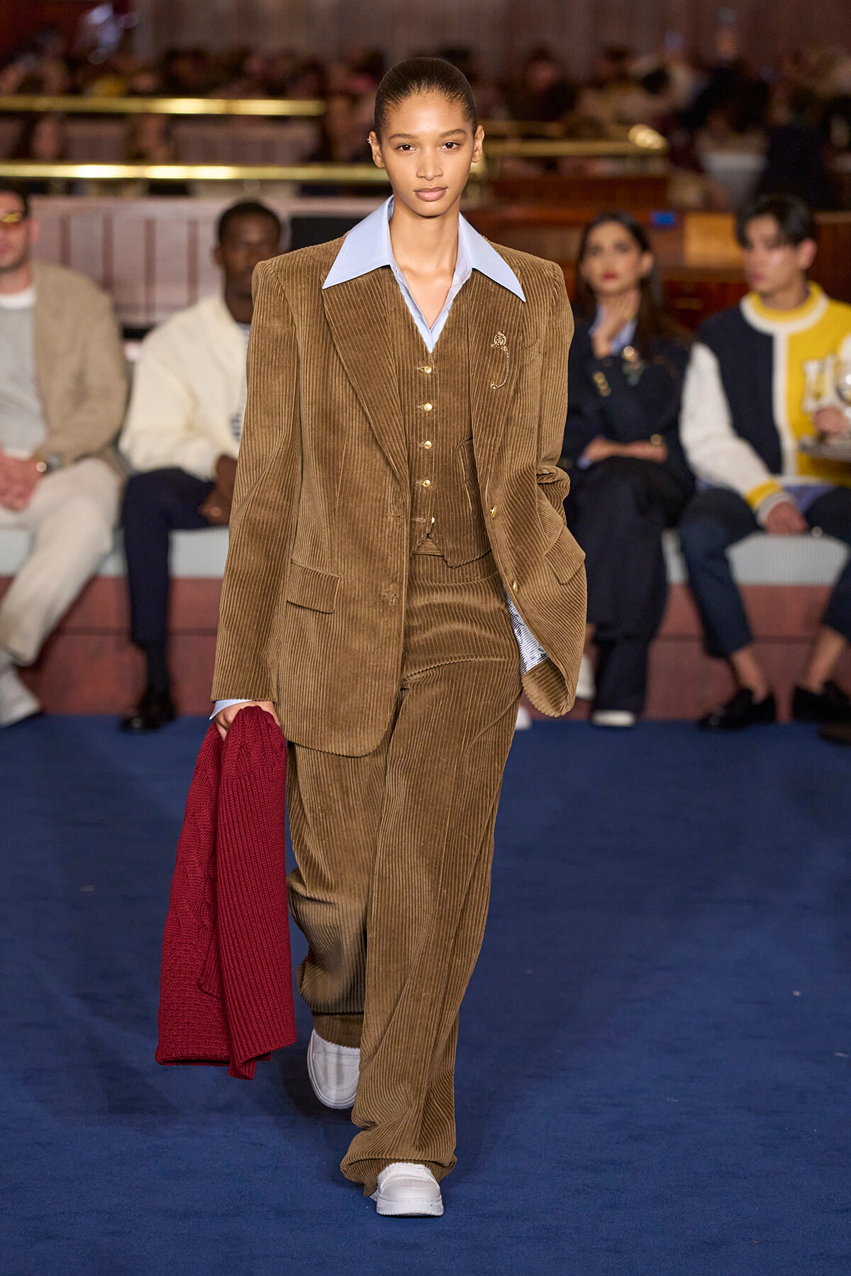 Model walking a fashion runway in a brown corduroy suit with a light blue shirt, holding a red cardigan in her left hand.