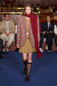 Model walking runway in a red scarf, beige pleated skirt, and plaid blazer.