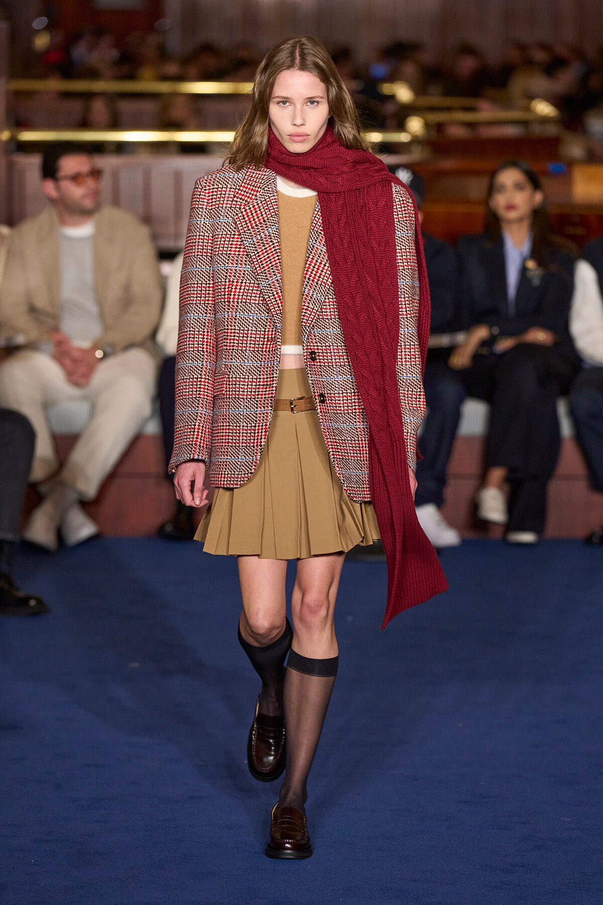 Model walking runway in a red scarf, beige pleated skirt, and plaid blazer.