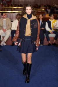 Model walks a fashion runway in a navy and tan varsity jacket, navy pleated skirt, black knee-high boots, and a small clutch.