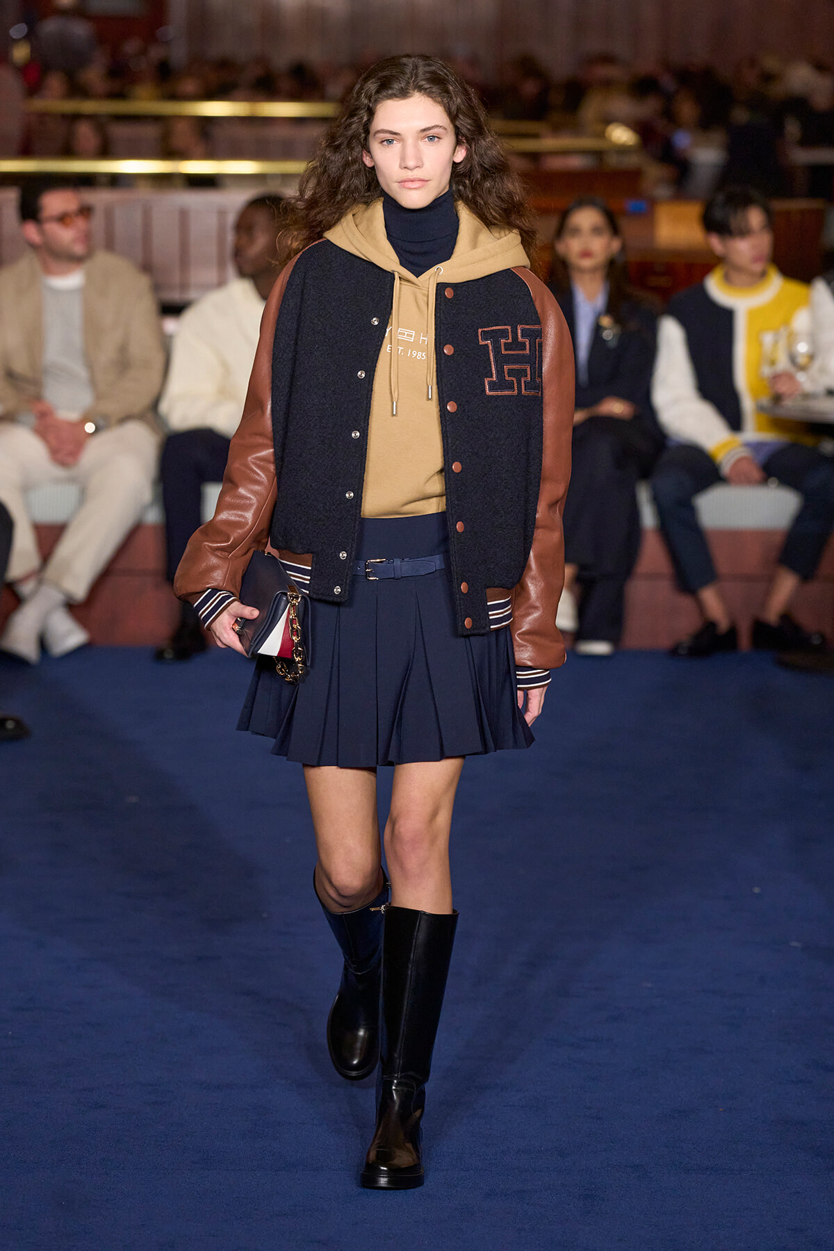 Model walks a fashion runway in a navy and tan varsity jacket, navy pleated skirt, black knee-high boots, and a small clutch.
