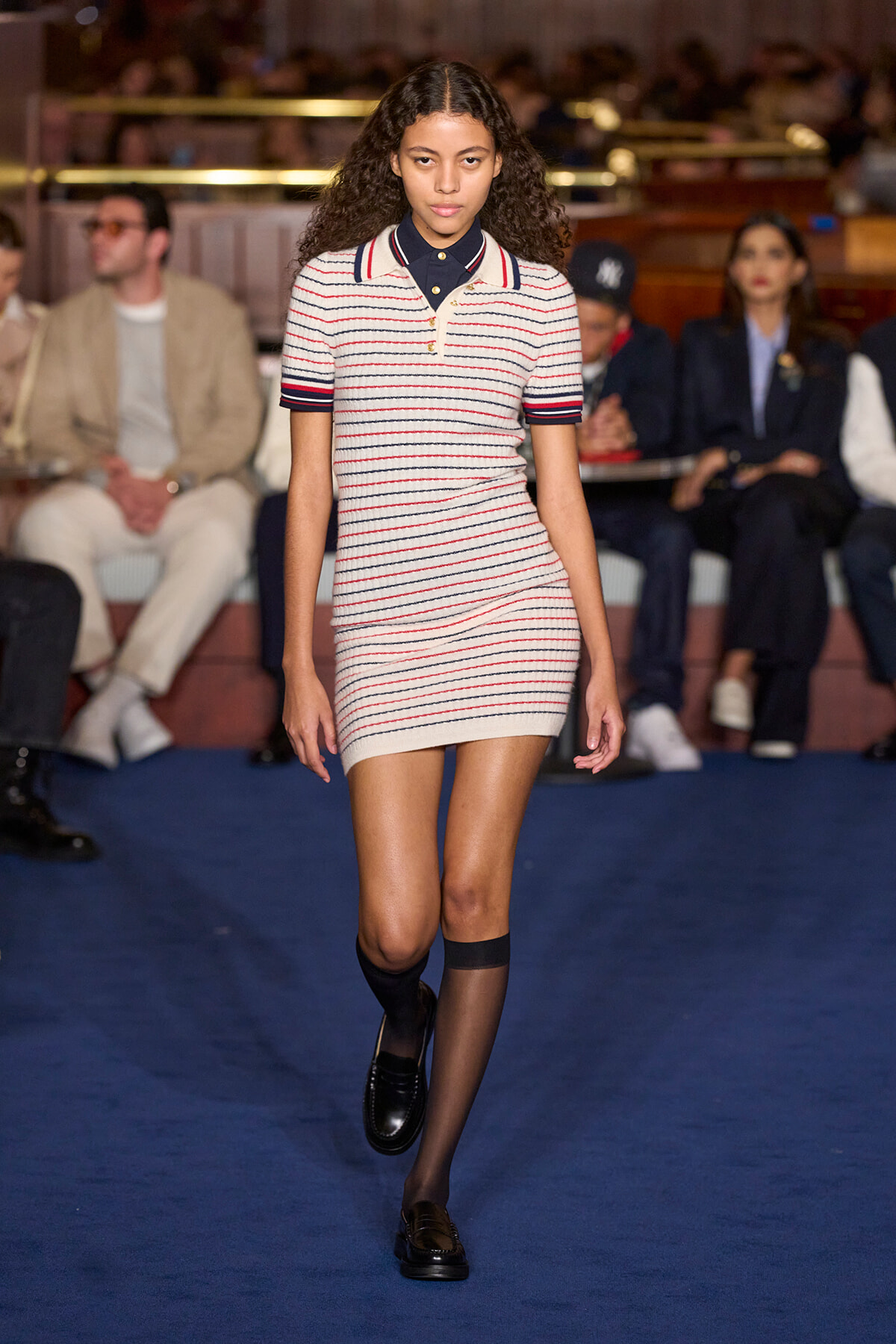 Model walks a runway wearing a cream-knit striped polo dress with navy and red stripes, navy collar, and black loafers; audience seated in background.