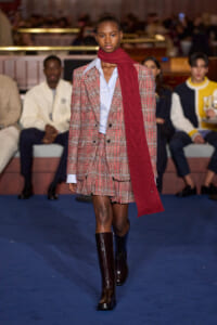 Model walks runway in red plaid blazer and matching skirt with a light blue shirt and burgundy scarf.