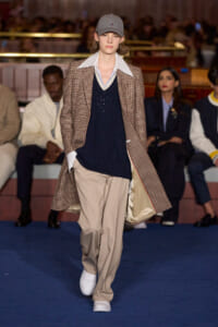 Runway model wearing a brown plaid coat over a navy sweater and beige trousers, with a gray cap and white sneakers.