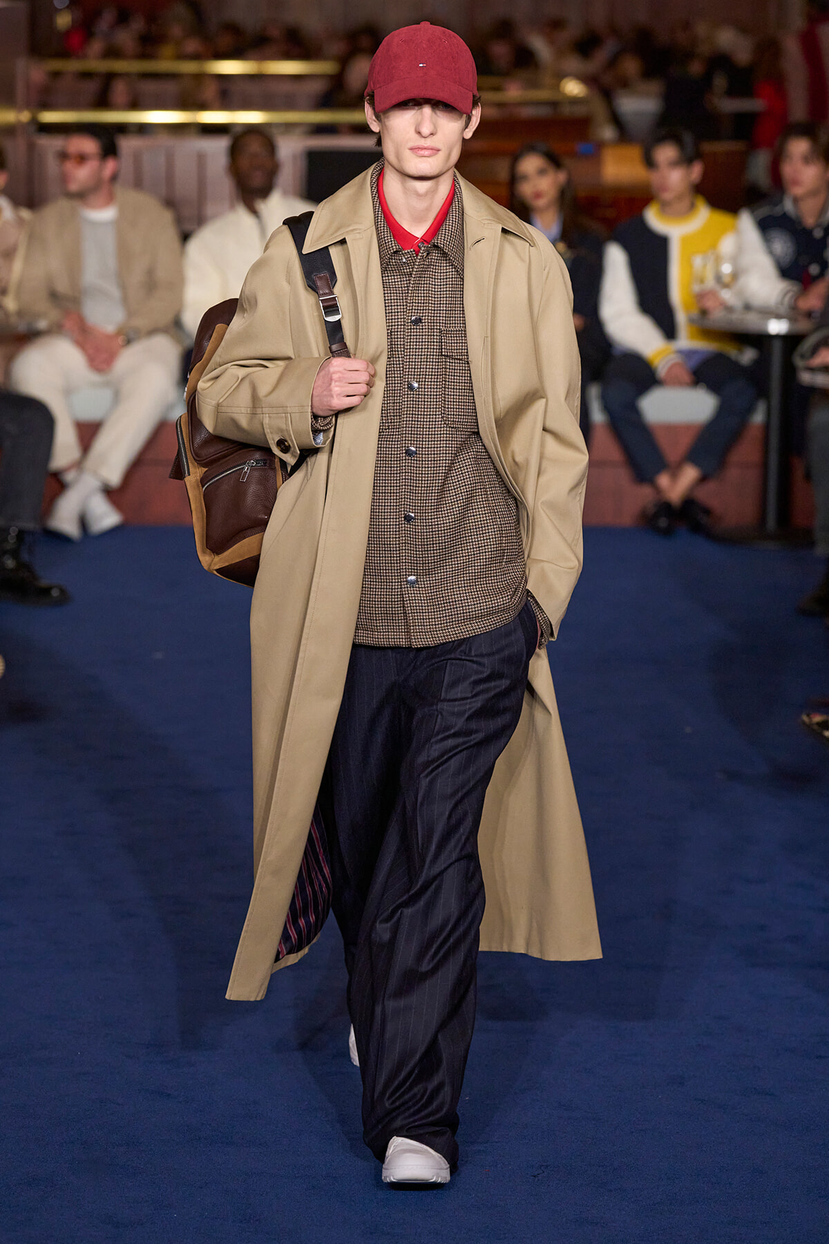 Male model on a runway wearing a beige trench coat over a brown checked shirt, red cap, dark trousers, white sneakers, and a brown backpack.