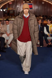 Male model on a runway wearing a tan cap, oversized brown plaid coat over a red knit sweater and beige pants; audience seated in the background.