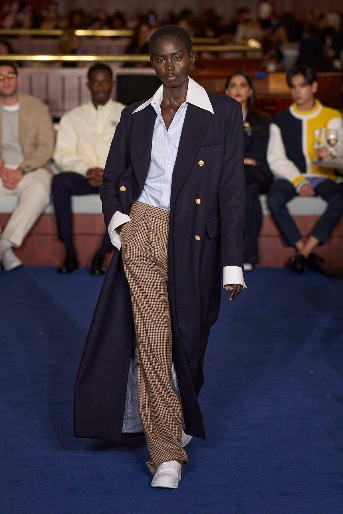 Model walking a runway in a navy double-breasted coat with gold buttons, light blue shirt, plaid brown trousers, and white sneakers.