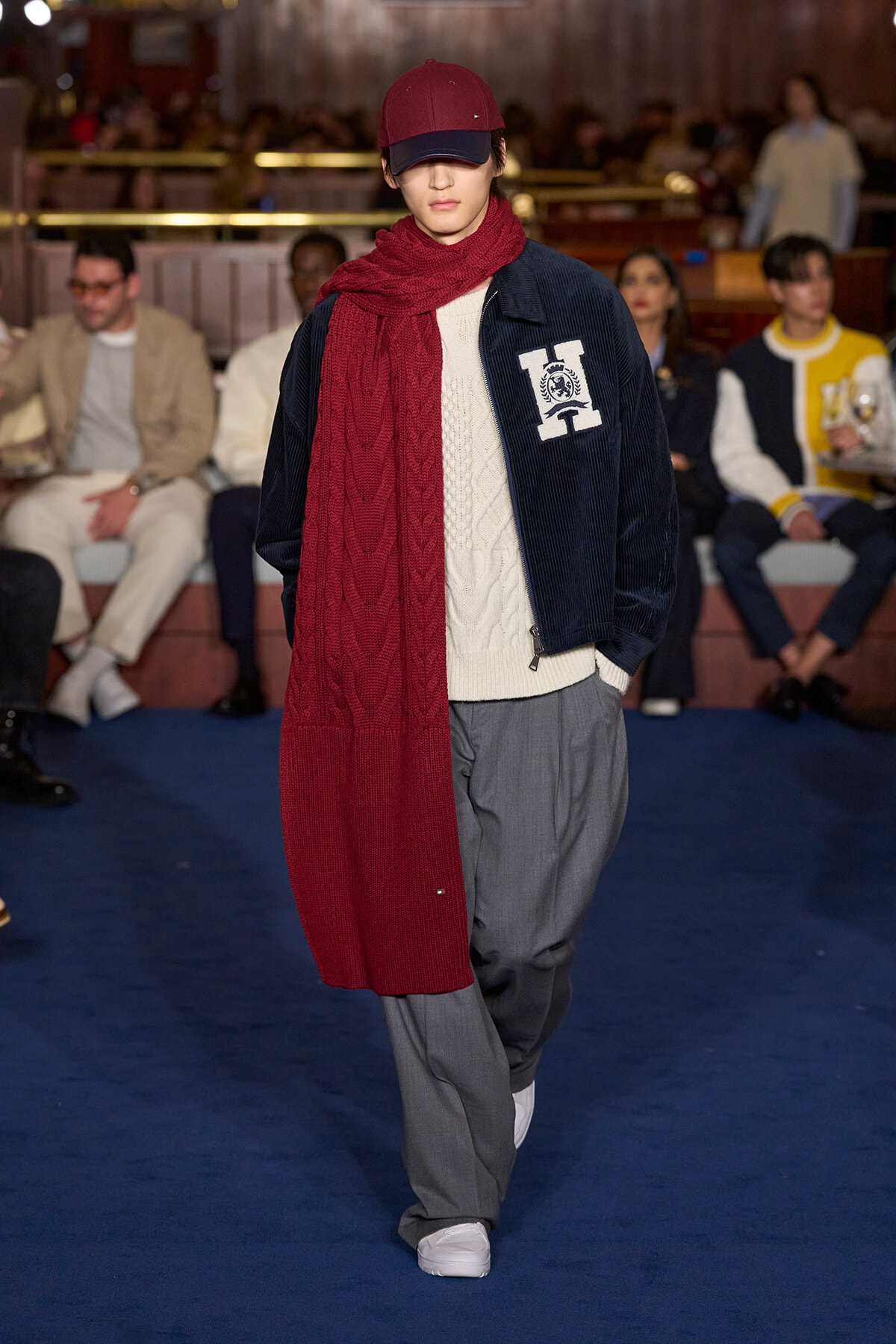 Model on runway wearing a burgundy scarf and cap with a navy corduroy jacket, cream sweater, and gray trousers on a blue runway surface.