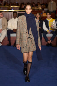 Model walks a fashion runway in a beige plaid blazer with a matching mini skirt, paired with a navy knit scarf and black ankle socks with loafers; audience seated along the catwalk in the background.