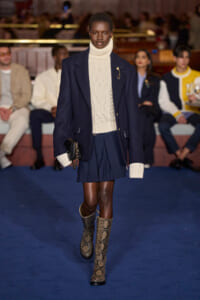 Female model walks the runway in a navy coat over a cream turtleneck sweater and navy pleated mini skirt; snakeskin boots and a small black clutch.