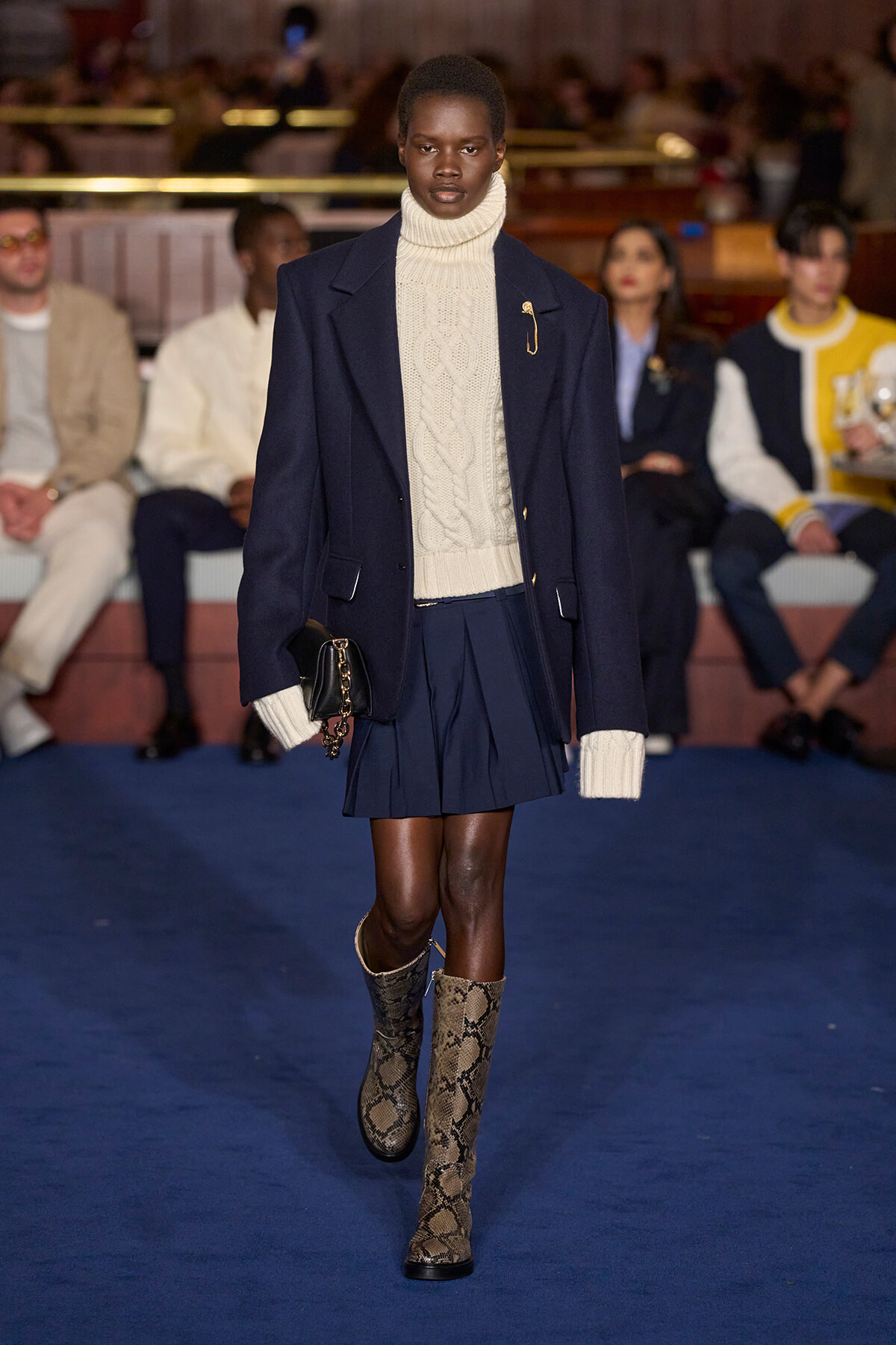 Female model walks the runway in a navy coat over a cream turtleneck sweater and navy pleated mini skirt; snakeskin boots and a small black clutch.
