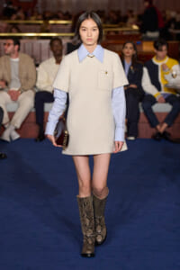 Fashion model walking a runway in a cream textured sheath with light blue shirt peeking at the collar; snakeskin boots, holding a dark clutch.