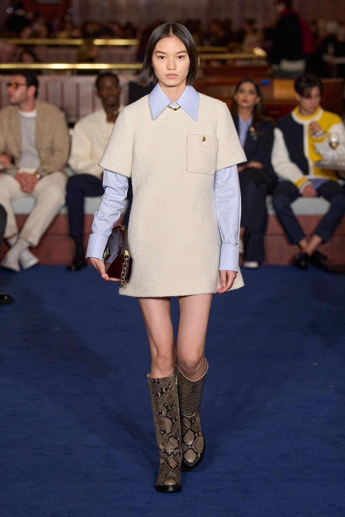 Fashion model walking a runway in a cream textured sheath with light blue shirt peeking at the collar; snakeskin boots, holding a dark clutch.
