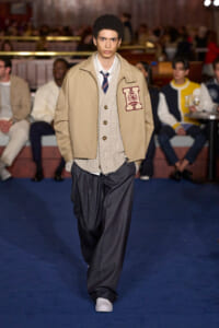 Male fashion model walking a runway in a beige jacket over a cream cardigan and striped tie, with dark trousers.