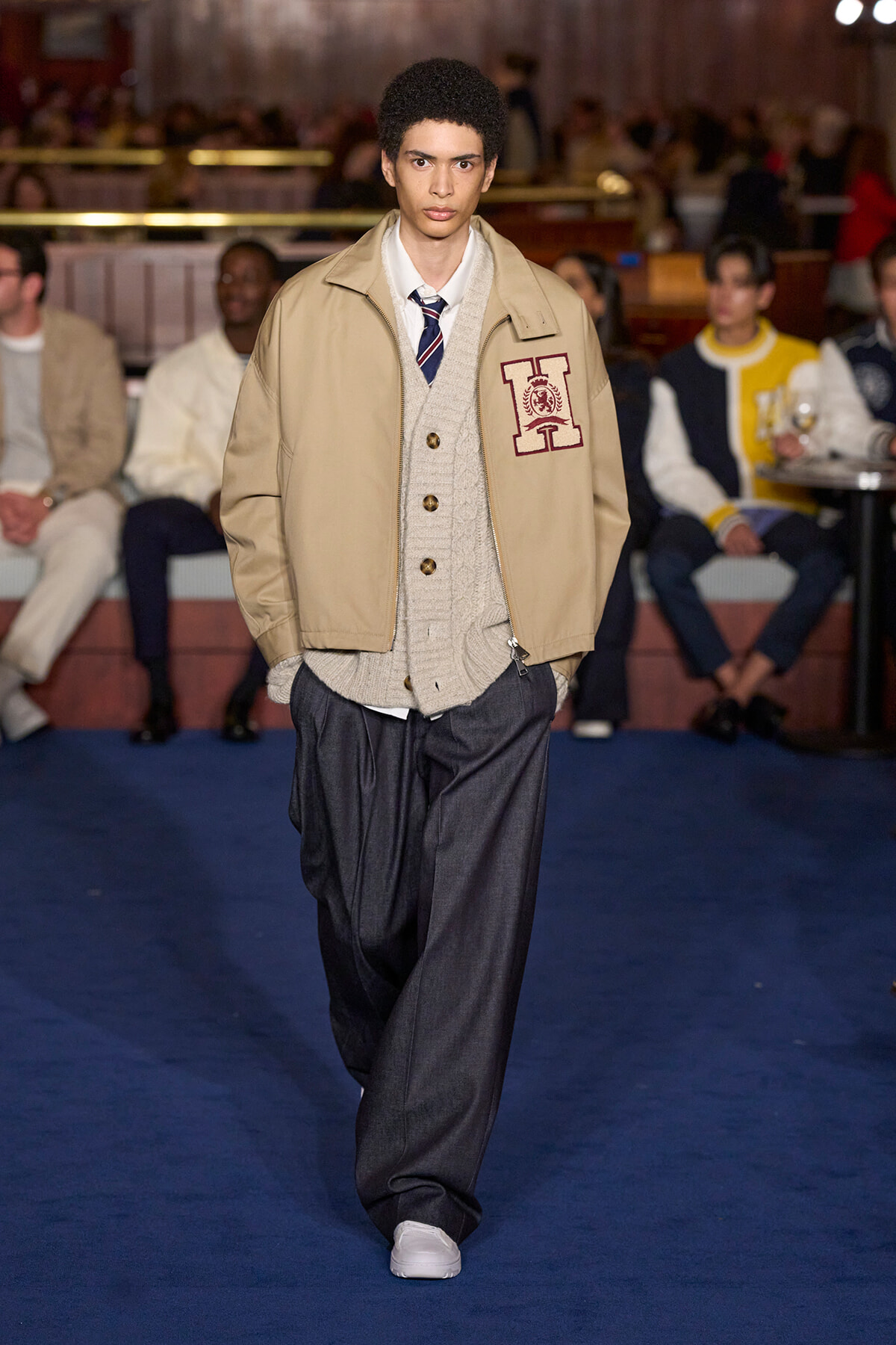 Male fashion model walking a runway in a beige jacket over a cream cardigan and striped tie, with dark trousers.