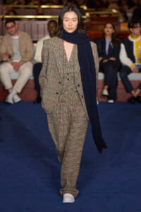 Model walking a fashion runway wearing a brown plaid suit with a navy scarf and white sneakers, on a blue carpeted runway.